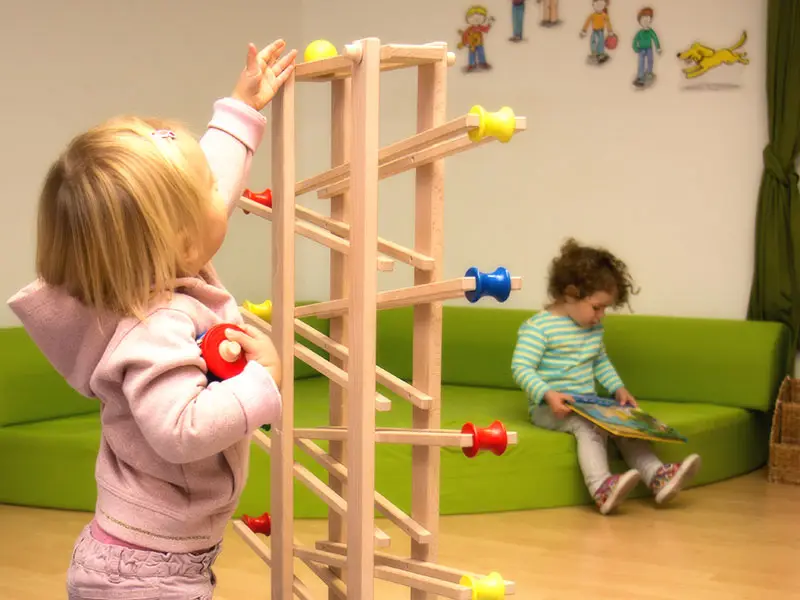 Toddler playing with a wooden ball ramp while another reads nearby