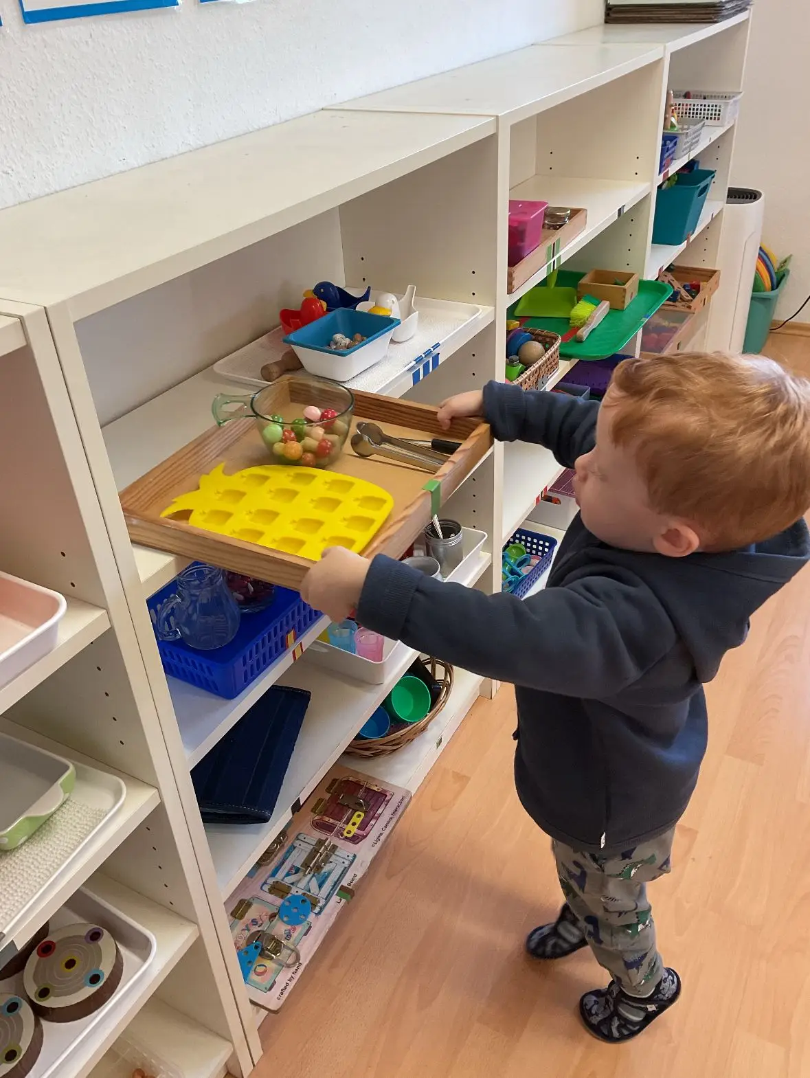 Toddler reaching for a Montessori practical life tray on a classroom shelf